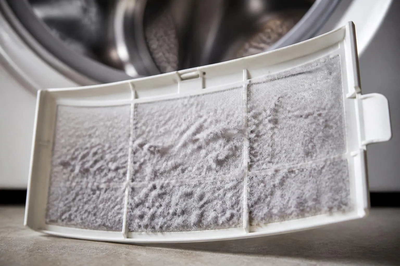 Filtre à peluches d'un sèche-linge sorti de son logement, tapis de fibres blanches sur la grille.